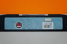 A rectangular object features a light blue panel with a stylized design and embedded graphics. On the left, a circular emblem displays a red hat with a white band and black background. On the right, a small, detailed graphic appears to depict a robot or mechanical figure in colors like blue and gold. Above the graphic, there's a label reading 'SINCE 2015'. The panel is bordered in a matte black frame with a vibrant orange background above.