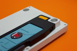 A close-up shot of a mechanical keyboard case with a unique keycap design. The background is orange, highlighting the device's metallic and plastic components. The keycaps feature distinct colors including light blue and black, with a small red triangle detail.