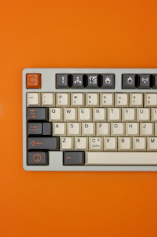 A mechanical keyboard is placed on a solid orange background. The keyboard features a mix of gray and cream-colored keys, with some keys having unique symbols, such as an exclamation point and various icons. The design blends vintage aesthetics with modern functionality.