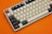 A mechanical keyboard with a mix of beige, black, and orange keys is set against an orange background. The keys feature unique and custom designs, including some with symbols instead of letters or numbers.
