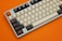A mechanical keyboard with a mix of beige, black, and orange keys is set against an orange background. The keys feature unique and custom designs, including some with symbols instead of letters or numbers.
