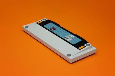 A gaming console cartridge with a predominantly white casing lies on an orange background. The cartridge includes a black and blue label area, with a small red circular detail in the center. A sticker featuring digital art is placed on the label area.