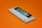 A gaming console cartridge with a predominantly white casing lies on an orange background. The cartridge includes a black and blue label area, with a small red circular detail in the center. A sticker featuring digital art is placed on the label area.