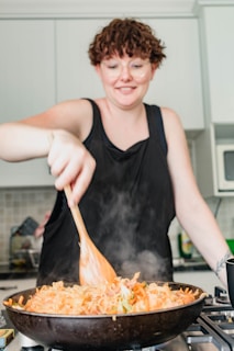 A person with curly hair is cooking in a modern kitchen, stirring a pan filled with steaming noodles and vegetables using a wooden spoon.