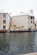 A waterfront building with beige exteriors and white shuttered windows stands adjacent to a calm water body. A seagull is in flight over the water, and there are potted plants near the building. The building features a sign reading 'La Caribe' and has balconies overlooking the water.