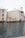 A waterfront building with beige exteriors and white shuttered windows stands adjacent to a calm water body. A seagull is in flight over the water, and there are potted plants near the building. The building features a sign reading 'La Caribe' and has balconies overlooking the water.