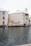 A waterfront building with beige exteriors and white shuttered windows stands adjacent to a calm water body. A seagull is in flight over the water, and there are potted plants near the building. The building features a sign reading 'La Caribe' and has balconies overlooking the water.
