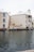A waterfront building with beige exteriors and white shuttered windows stands adjacent to a calm water body. A seagull is in flight over the water, and there are potted plants near the building. The building features a sign reading 'La Caribe' and has balconies overlooking the water.