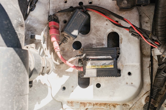 A car engine compartment shows electrical components, including a module with wiring. The wires are red and black, connected to a rectangular black device, and the metal surface underneath is slightly rusted and dirty.