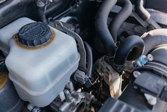 A close-up view of a car engine compartment, featuring a coolant reservoir with a black cap, various hoses, pipes, and metal components. The lighting casts natural highlights and shadows on the components.