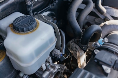 A close-up view of a car engine compartment, featuring a coolant reservoir with a black cap, various hoses, pipes, and metal components. The lighting casts natural highlights and shadows on the components.