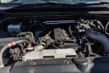 Close-up view of a car engine compartment with various components such as pipes, connectors, and an engine block. The setting is well-lit, showcasing different parts made of metal and plastic.