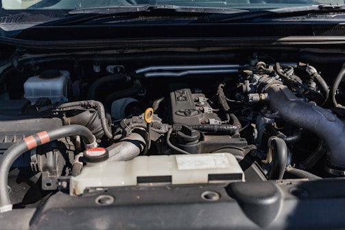 Close-up view of a car engine compartment with various components such as pipes, connectors, and an engine block. The setting is well-lit, showcasing different parts made of metal and plastic.