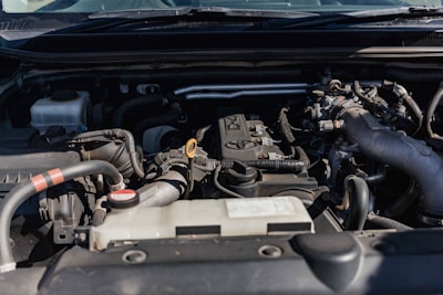 Close-up view of a car engine compartment with various components such as pipes, connectors, and an engine block. The setting is well-lit, showcasing different parts made of metal and plastic.