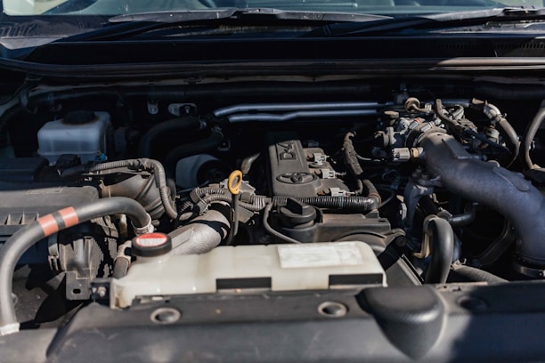Close-up view of a car engine compartment with various components such as pipes, connectors, and an engine block. The setting is well-lit, showcasing different parts made of metal and plastic.
