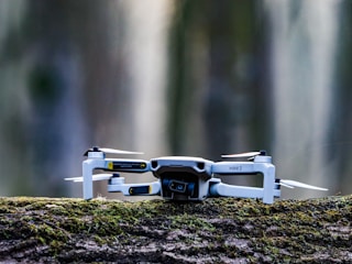 A small drone with a camera, labeled 'MINI 2', is placed on a moss-covered log. The background is blurred, giving a sense of depth and focus on the drone itself.