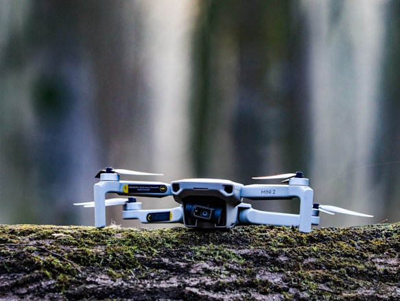 A small drone with a camera, labeled 'MINI 2', is placed on a moss-covered log. The background is blurred, giving a sense of depth and focus on the drone itself.
