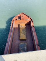 Aerial view of a large cargo ship's deck with rusted surfaces and two prominent, weathered steel plates. The vessel is surrounded by teal green water under clear daylight.