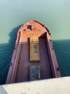 Aerial view of a large cargo ship's deck with rusted surfaces and two prominent, weathered steel plates. The vessel is surrounded by teal green water under clear daylight.