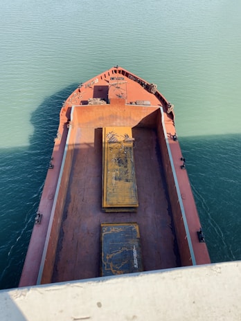 Aerial view of a large cargo ship's deck with rusted surfaces and two prominent, weathered steel plates. The vessel is surrounded by teal green water under clear daylight.