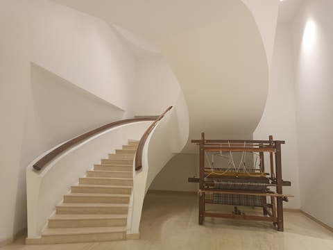A minimalist, well-lit interior space features a curved staircase with a smooth, white finish and dark wood railing. Adjacent to the staircase, a traditional wooden loom with partially woven fabric is present, adding a touch of craftsmanship to the contemporary setting.