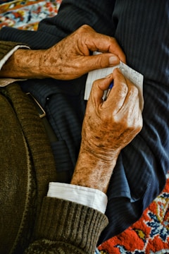 An elderly person's hands are holding a small piece of paper, which appears to have notes or writing on it, and a pen. The person is wearing a sweater and a white shirt underneath. The background includes colorful, patterned fabric.