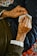 An elderly person's hands are holding a small piece of paper, which appears to have notes or writing on it, and a pen. The person is wearing a sweater and a white shirt underneath. The background includes colorful, patterned fabric.