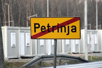 A road sign with the name Petrinja, featuring a red diagonal line crossing through it. Behind the sign, there are several portable buildings or containers. The background includes a wooded area with leafless trees.