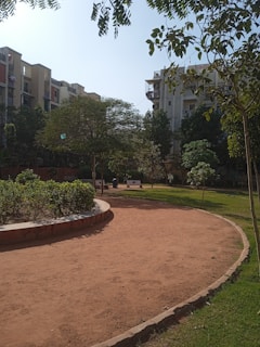 A residential area featuring apartment buildings in the background, with a small park in the foreground. There are trees, shrubs, and benches along a dirt path, and the scene is bathed in natural daylight.