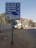 A blue directional road sign features names and directions in both Hindi and English. Behind the sign, a busy urban street is lined with high-rise buildings and a few trees. A strip of sandy sidewalk with some litter runs parallel to the road. The background includes some parked vehicles and a person walking, indicative of an active city street environment.