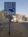 A blue directional road sign features names and directions in both Hindi and English. Behind the sign, a busy urban street is lined with high-rise buildings and a few trees. A strip of sandy sidewalk with some litter runs parallel to the road. The background includes some parked vehicles and a person walking, indicative of an active city street environment.