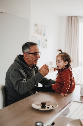 An older man wearing glasses and a grey hoodie is feeding a toddler in a red sweater sitting on a kitchen counter. The man is holding out a spoon to the child, who is about to eat from it. A plate with some food remains is on the counter in front of them. The background includes a white wall with picture frames and a window with beige curtains allowing natural light to fill the room.