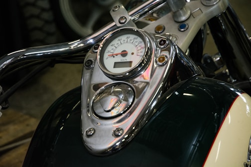 A close-up view of a motorcycle's instrument panel featuring a speedometer and several control buttons. The chrome details and handlebars reflect light, adding a polished look to the vehicle. The background shows part of a tire and floor.