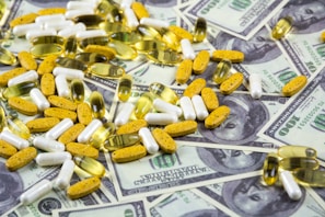 A collection of various pills and capsules, including yellow, white, and transparent ones, is scattered over a background of hundred-dollar bills.