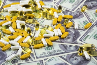 A collection of various pills and capsules, including yellow, white, and transparent ones, is scattered over a background of hundred-dollar bills.