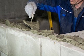 A person wearing gloves is applying mortar on top of cinder blocks using a trowel. The individual is dressed in a blue work jacket. The background includes a partially visible yellow ladder and a gray concrete wall.