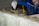 A person wearing gloves is applying mortar on top of cinder blocks using a trowel. The individual is dressed in a blue work jacket. The background includes a partially visible yellow ladder and a gray concrete wall.