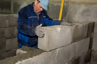 A worker in blue overalls and a cap is placing a large cinder block onto a row of similar blocks, constructing a wall. He is wearing gloves and appears focused on aligning the block correctly, with a yellow spirit level in the background for accuracy.