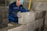 A worker in blue overalls and a cap is placing a large cinder block onto a row of similar blocks, constructing a wall. He is wearing gloves and appears focused on aligning the block correctly, with a yellow spirit level in the background for accuracy.