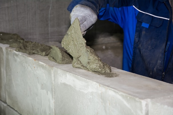 A worker is applying mortar onto a masonry wall. The person is wearing a blue uniform and a protective glove, using a trowel to spread the mortar.
