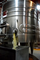 A large stainless steel fermentation tank is positioned prominently, with liquid visibly flowing from a tap or spout into a rectangular container below. The tank has a glossy surface, reflecting light, and features multiple riveted seams. There is a sign with a number '43' attached to the upper section of the tank.
