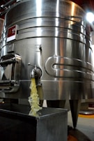 A large stainless steel fermentation tank is positioned prominently, with liquid visibly flowing from a tap or spout into a rectangular container below. The tank has a glossy surface, reflecting light, and features multiple riveted seams. There is a sign with a number '43' attached to the upper section of the tank.