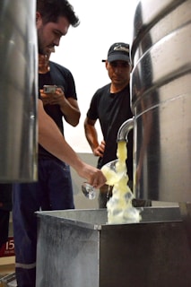 Three people are gathered around a large stainless steel container, observing as a liquid pours from a spout into a glass held by one of them. The setting appears industrial, likely inside a brewery or similar facility.