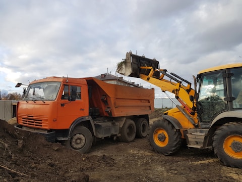 A large orange dump truck is being loaded with dirt by a yellow backhoe loader on a construction site. The vehicles are situated on a muddy surface, and the background shows a cloudy sky with muted colors.