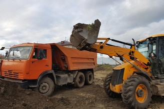An orange dump truck and a yellow backhoe loader are engaged in a construction activity. The backhoe loader is transferring a bucket of soil and debris into the dump truck. The site appears to be a construction area with dirt and some buildings in the background under a cloudy sky.