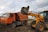 An orange dump truck and a yellow backhoe loader are engaged in a construction activity. The backhoe loader is transferring a bucket of soil and debris into the dump truck. The site appears to be a construction area with dirt and some buildings in the background under a cloudy sky.