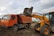 An orange dump truck and a yellow backhoe loader are engaged in a construction activity. The backhoe loader is transferring a bucket of soil and debris into the dump truck. The site appears to be a construction area with dirt and some buildings in the background under a cloudy sky.