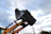 Close-up of a rugged excavator bucket digging into rocky soil under a bright blue sky.
