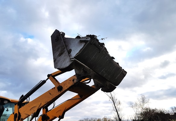 A large metal excavator bucket attached to a yellow arm extending upwards against a cloudy sky backdrop. The bucket appears to be lifting soil or debris, as some material is visible along its edge. A portion of the vehicle cabin is visible in the bottom left corner.