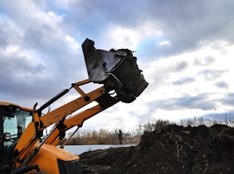 A construction vehicle with an articulated arm and a large metal scoop is featured prominently, raised against a backdrop of a cloudy sky. The vehicle is positioned on uneven, earthy terrain with some sparse vegetation visible. The vehicle is primarily orange, and the scene suggests a construction or excavation site.
